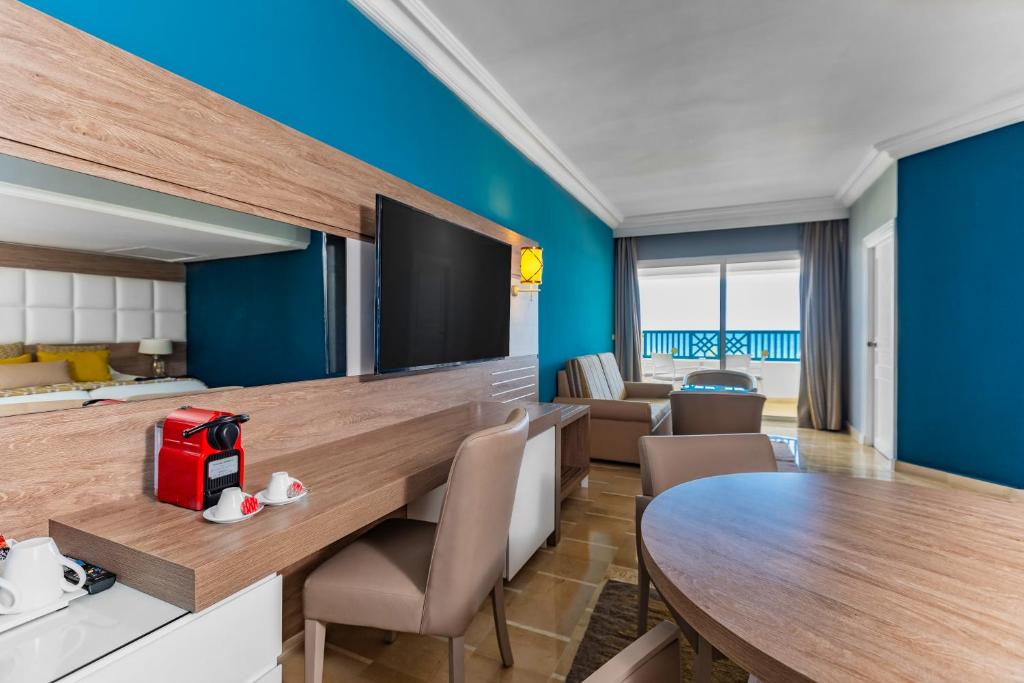 Mūsdienīgs viesnīcas numurs ar zilu sienu, koka mēbelēm un plašu logu, kas sniedz skatu uz terasi. Uz galda atrodas kafijas automāts un trīs kafijas krūzītes.