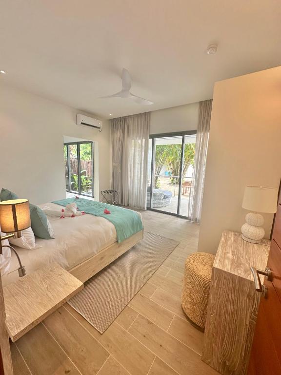 Mūsdienīga guļamistaba ar gaisīgu interjeru, kas ietver plašu logu ar skatu uz āra terasi. Gultā ir zila sega un dekoratīvi spilveni, bet blakus atrodas koka naktsgaldiņš un lampiņa.