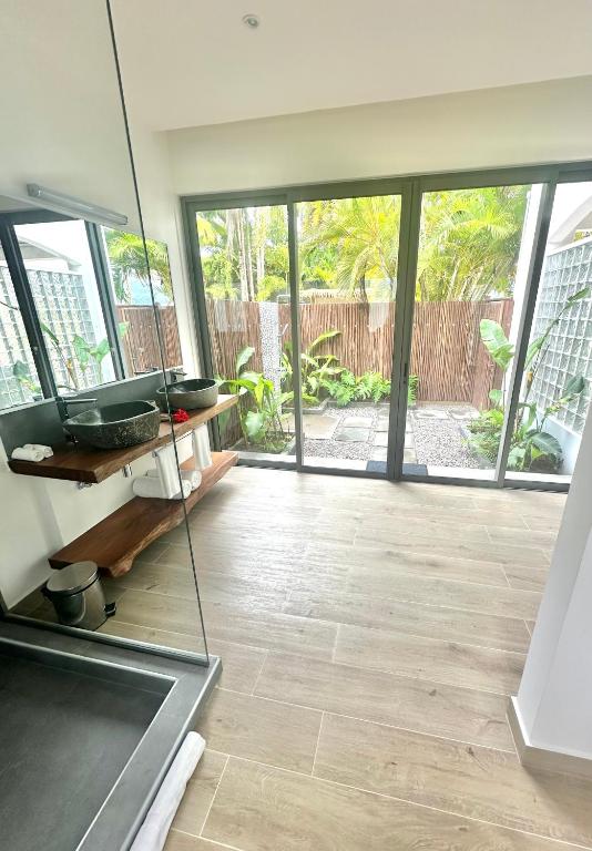 Mūsdienīgs vannas istabas interjers ar stikla sienām un skatu uz zaļo dārzu. Uz koka plaukta atrodas akmens izlietne un dvieļi, bet grīda ir no koka.