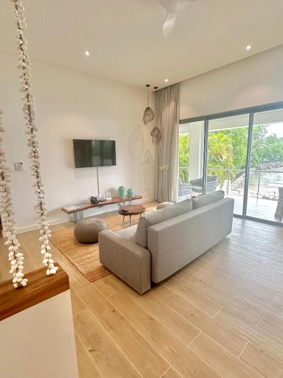 Mūsdienīgs viesistaba ar pelēku dīvānu, televizoru uz sienas un lieliem logiem, kas nodrošina dabīgu apgaismojumu un skatu uz āra terasi. Interjers ir vienkāršs un elegants, ar dabīgiem materiāliem un neitrālām krāsām.