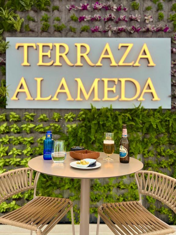 Terasa ar nosaukumu "Terraza Alameda", kur uz galda redzami dzērieni un uzkodas, fons ar zaļām augiem.