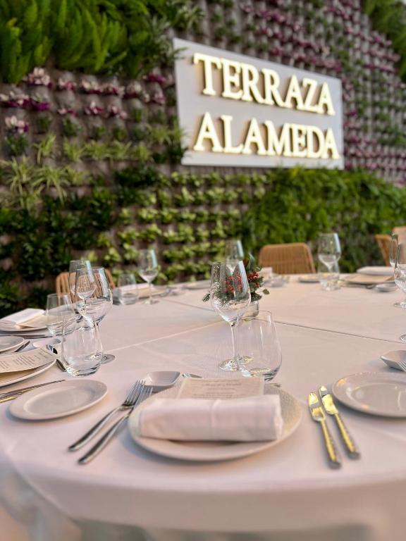 Elegantā restorāna "Terraza Alameda" galda iestatījums ar baltu galdautu, stikla traukiem un augu sienu fonā. Izsmalcināta atmosfēra, kas paredzēta īpašām svinībām.
