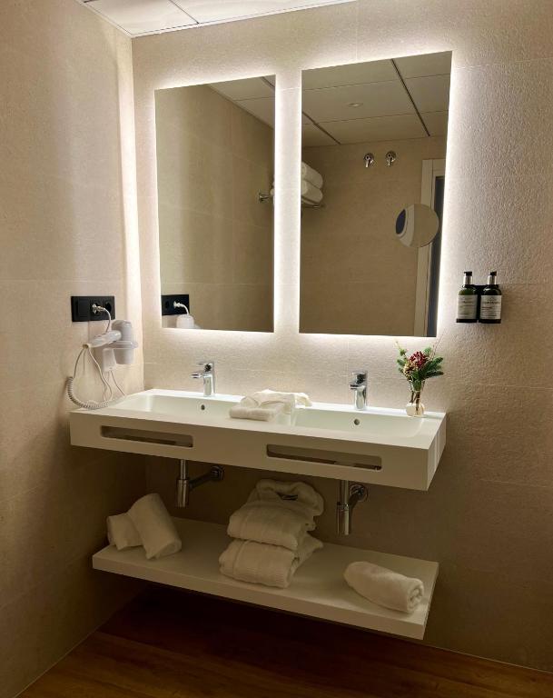 Mūsdienīgs vannas istabas dizains ar diviem spoguļiem, divām izlietnēm un sakārtotiem dvieļiem uz plaukta. Telpa ir gaiša un stilīga, piedāvājot komfortablu vidi.