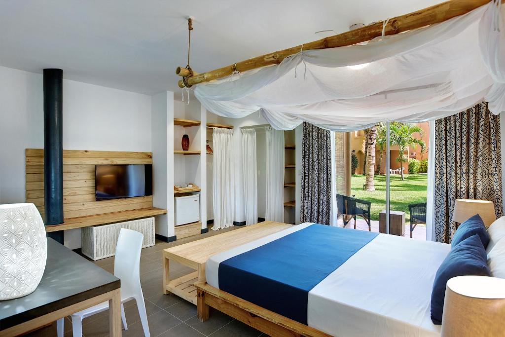 Mūsdienīga guļamistaba ar koka mēbelēm, gultu ar baltu drānu un skatu uz dārzu. Telpā ir televizors un galds, kas radītu relaksējošu atmosfēru.