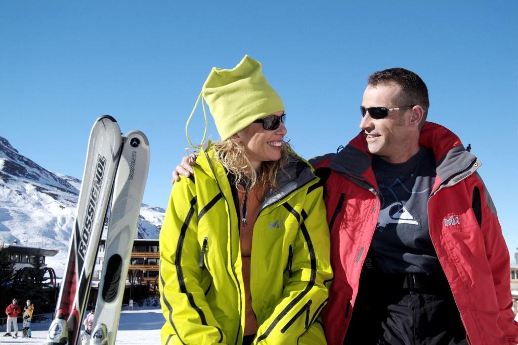 Divi cilvēki, tērpti krāsainā slēpošanas apģērbā, priecīgi sarunājas kalnu vidē, fonā redzami sniegoti kalni un skijiem.