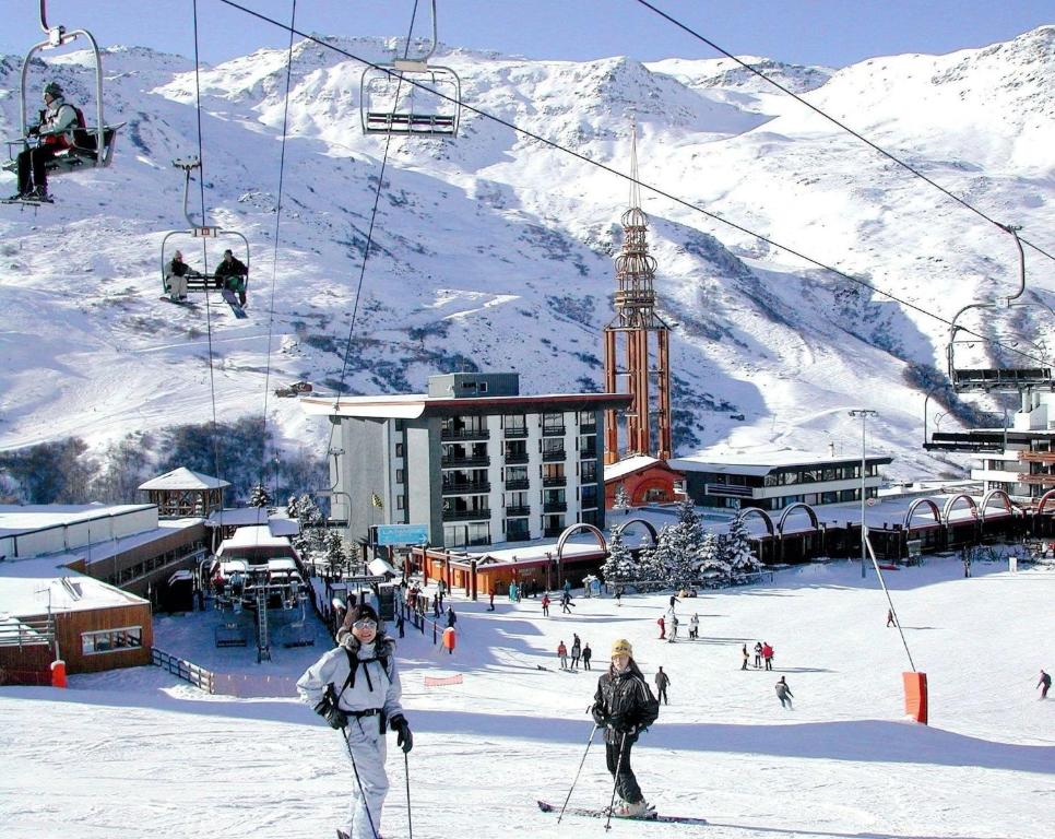 Sniega kūrorts ar slēpotājiem, slēpošanas pacēlājiem un kalniem fona. Apkaimē redzams viesnīcu komplekss un slēpošanas zonas aktivitātes.