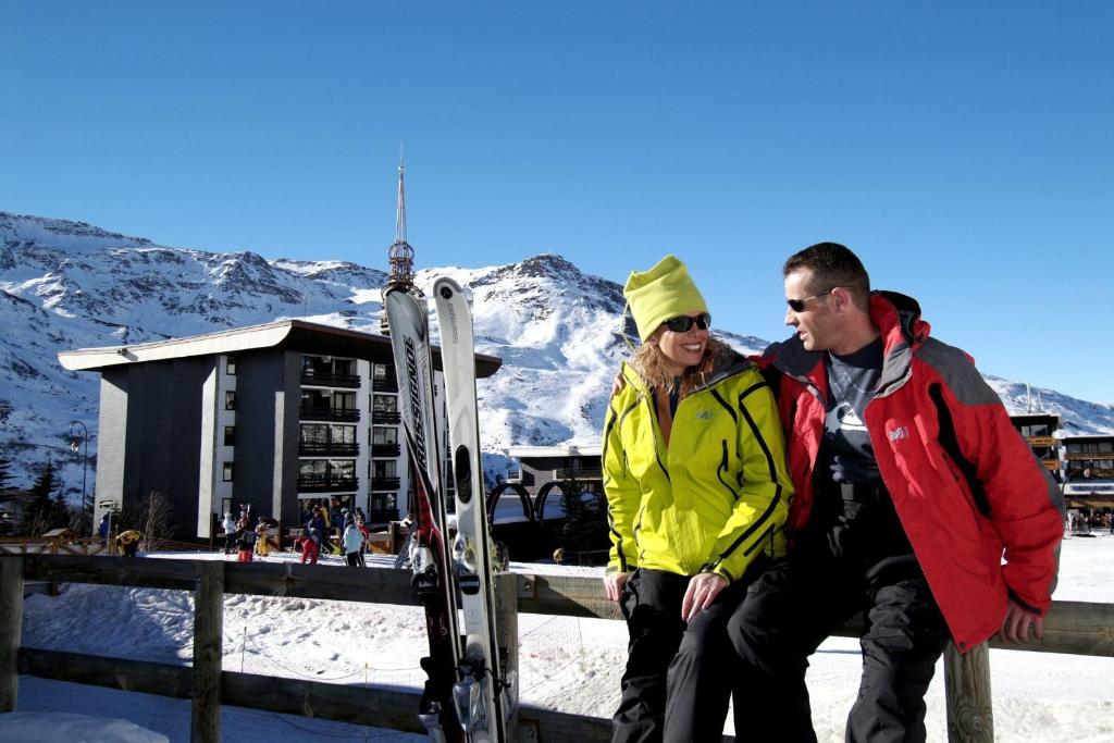 Sniega slēpošanas kūrorts ar pāri, kas baudīja sauli un ziemas aktivitātes. Fons ar kalniem un viesnīcu, kas piepildīts ar slēpotājiem.