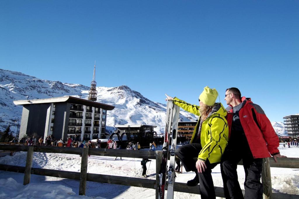 Divi slēpotāji priecājas par kalnu ainavu, skatoties uz apkārtni, kamēr fons ir sniegotā kalnu ainava un viesnīcas.