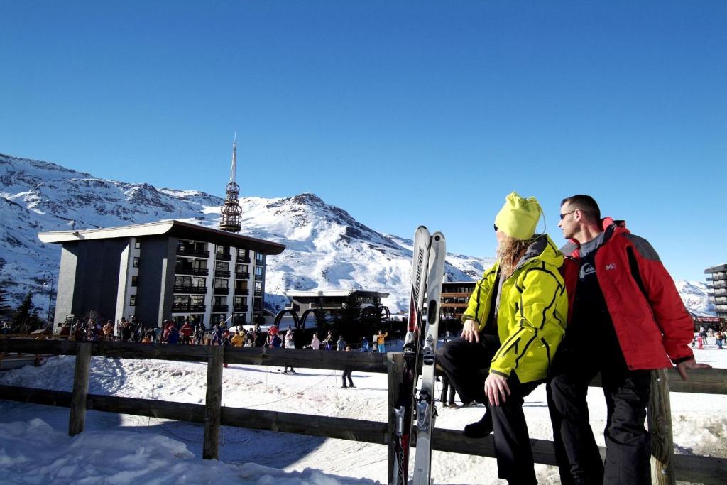 Pāris, kas tērpies ziemas apģērbā, atpūšas pie slēpošanas kūrorta, kamēr sniegots kalns un skaidra debesis veido skaistu fonu. Sniega segums un cilvēki slēpošanas trasē piešķir dzīvīgumu ainai.