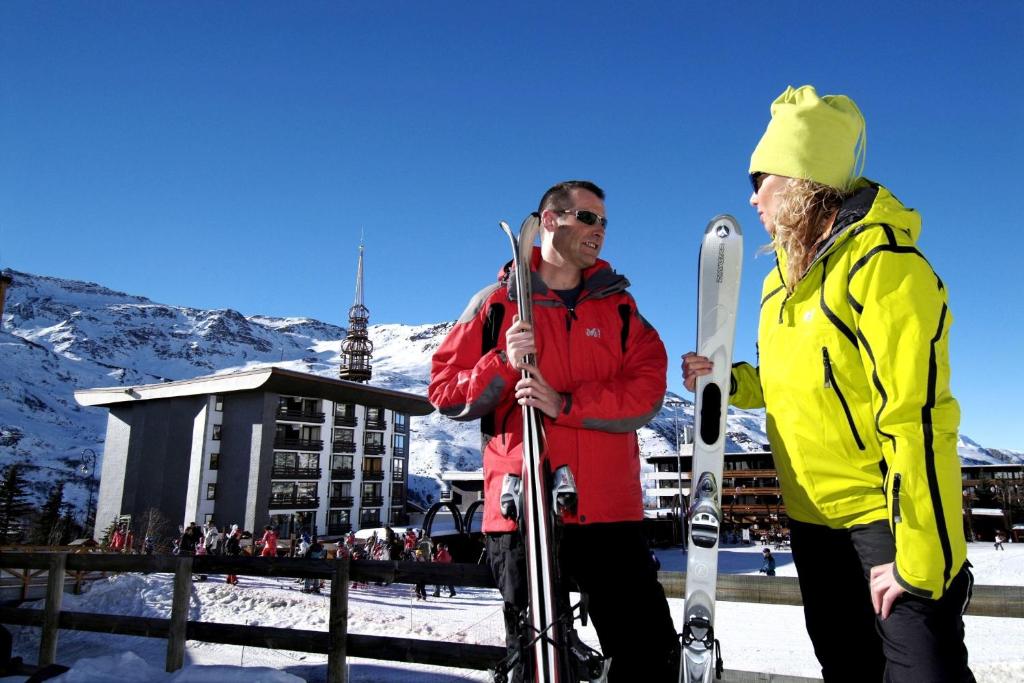Slēpošanas entuziasti sarunājas kalnu kūrortā, tērpti siltos ziemas apģērbos, ar slēpēm rokās. Fons veido sniegots kalns un skaidrs zils debesis.
