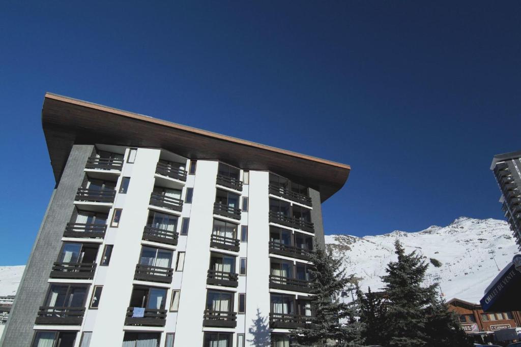 Mūsdienīgs kalnu viesnīcas ēka ar koka jumtu, ko ieskauj sniegoti kalni un skaidrs zils debesis.