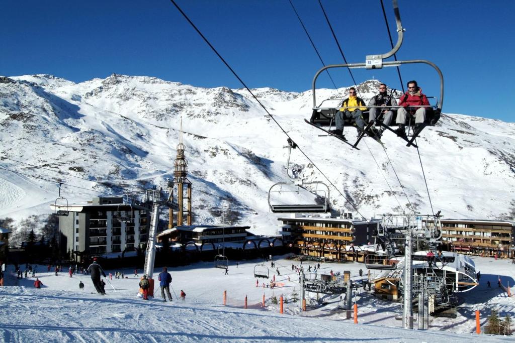 Sniegotā kalnu ainava ar slēpotājiem un sniega pacēlāju, kas ved uz augšu. Fons ietver viesnīcu un slēpošanas infrastruktūru.