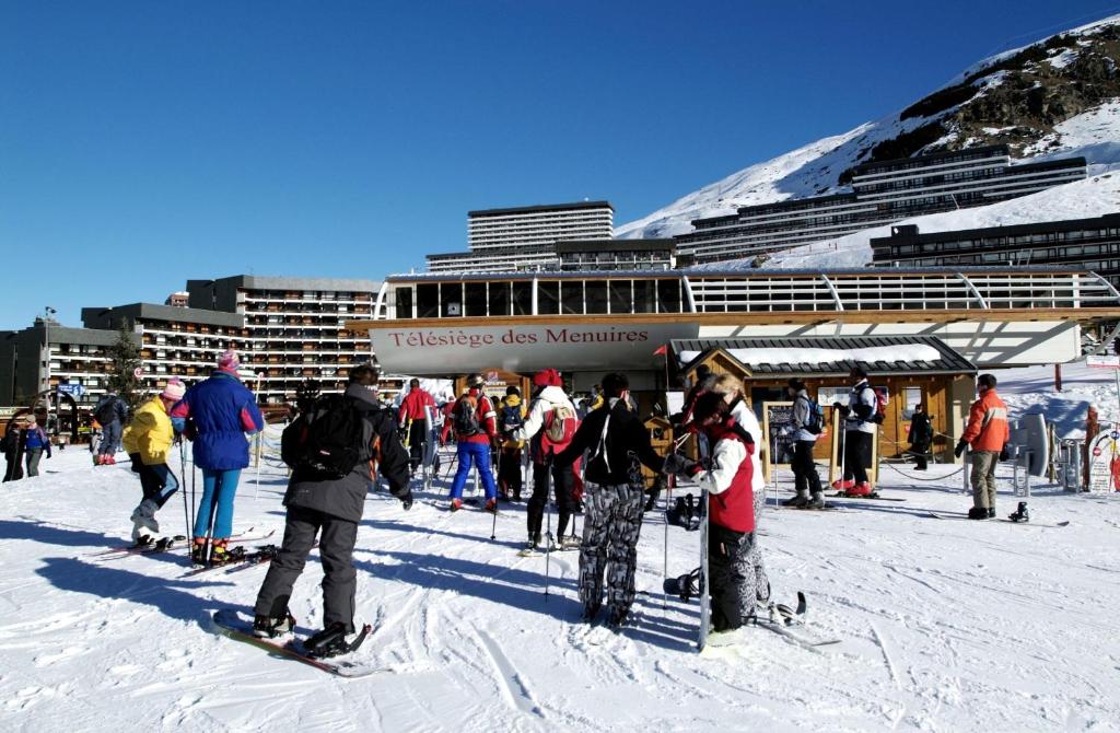 Slēpotāju grupas pulcējas pie pacēlāja "Téléskiège des Menuires", kamēr fonā redzamas kalnu viesnīcas un skaidrs zils debesis.