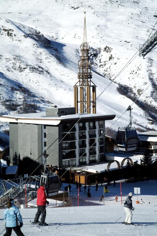 Ziemas sporta kūrorts ar slēpotājiem sniegotā ainavā, fonā redzama moderna ēka un gaisa tramvajs.