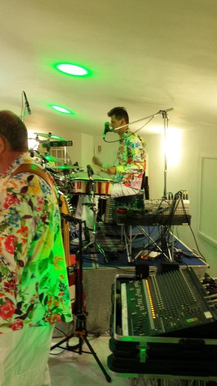 Mūziķis spēlē bungas un dzied mikrofonā, tērpies krāsainā ziedu rakstā, kamēr fons ir apgaismots ar zaļām gaismām. Aiz viņa redzami skaņu aprīkojumi un citi instrumenti.