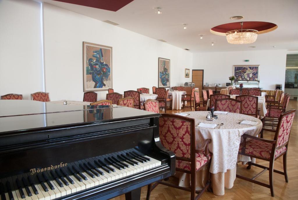 Elegants restorāns ar melnu klavieri priekšplānā, rotāts ar greznām krēslām un galdiem. Sienas dekorētas ar mākslas darbiem, radot patīkamu atmosfēru.