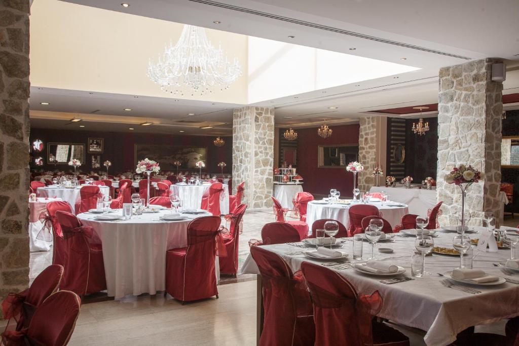 Elegants restorāns ar apaļām galdiem, kas klāti baltos galdautos un sarkanās krēslā. Telpā ir ierīkoti kristāla lustri un ziedu dekorācijas, radot svinīgu atmosfēru.