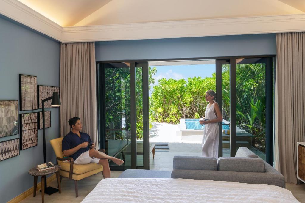 Mūsdienīgas viesnīcas numura iekārtojums ar skatu uz dārzu. Vīrietis sēž krēslā, bet sieviete stāv pie durvīm, turot dzērienu.