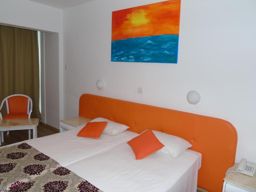 Gaišas viesnīcas numurs ar divguļamo gultu un oranžu galvgali, dekorēts ar krāsainu saulrietā gleznu. Blakus atrodas balta krēsla un galda komplekts.