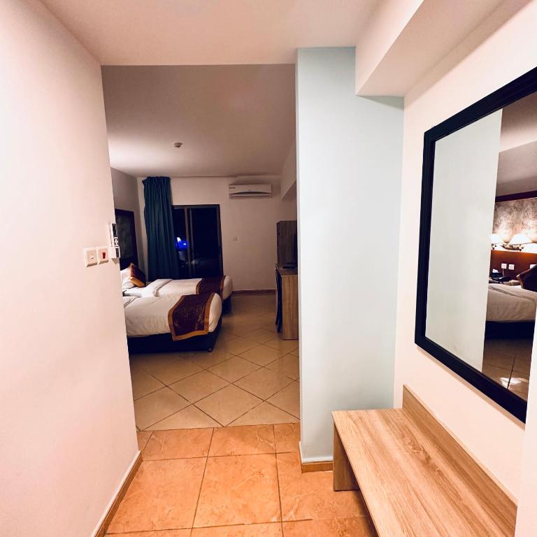 Mūsdienīga viesnīcas numura ieeja ar divām gultām, televizoru un gaisīgu interjeru. Attēlā redzama arī spoguļa un koka mēbeļu daļa.