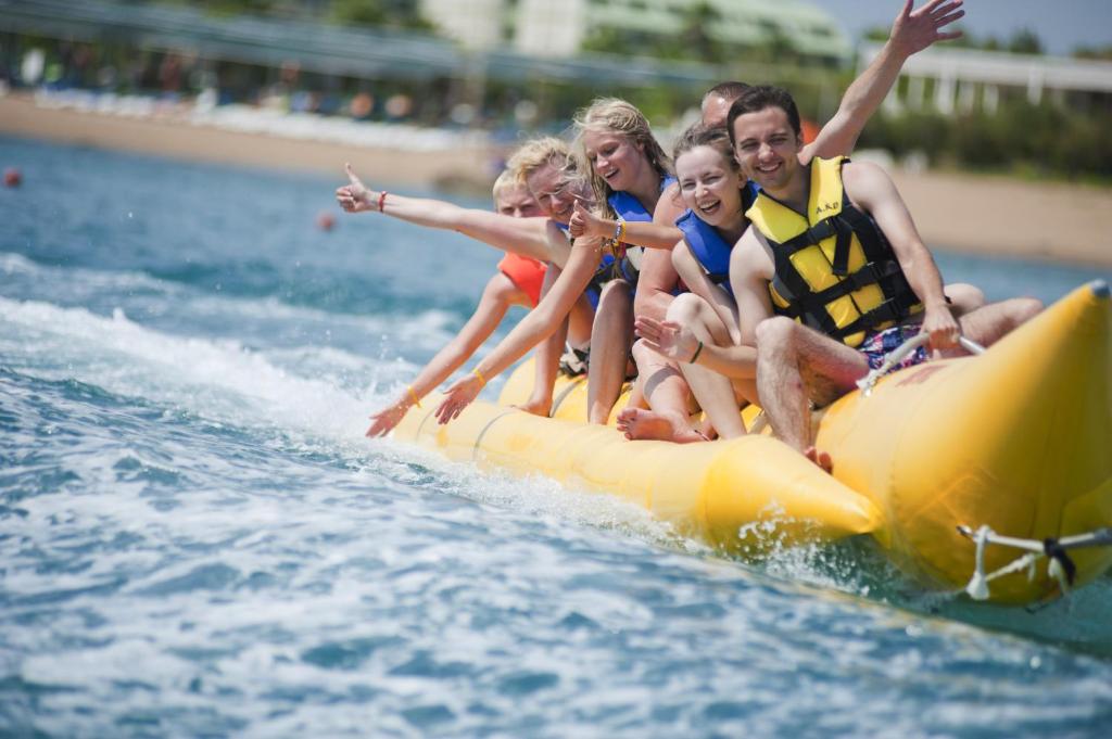 Priecīgi cilvēki brauc ar banānu laivu pa ūdeni, izsaucot prieka saucienus un baudot vasaras atpūtu. Dzeltenā laiva ir piepildīta ar sešiem cilvēkiem, kas valkā glābšanas vestes.