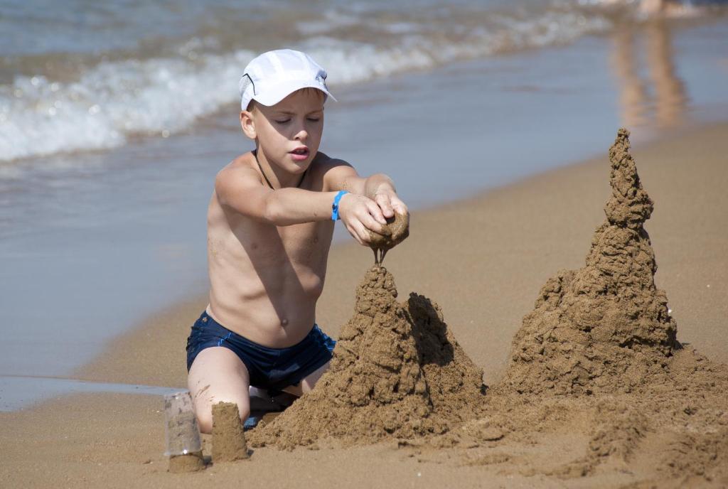 Bērns pludmalē būvē smilšu pili, koncentrējoties uz detaļām. Apkārt ir jūra un saulains laiks.