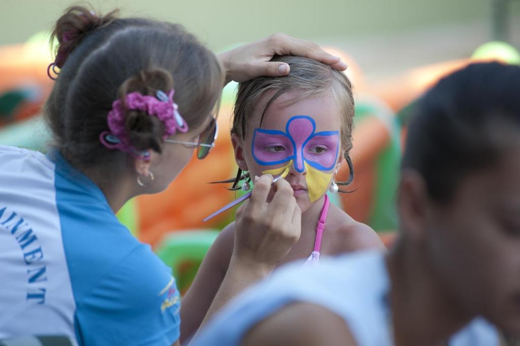 Bērns ar krāsotu seju saņem grimu no mākslinieces, kas uzmanīgi izmanto otu. Fons ir izklaides vieta, kurā redzami krāsaini elementu.