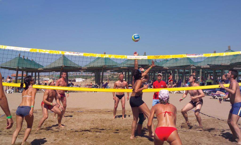 Pludmales volejbola spēle, kurā piedalās vairākas personas, izbaudot saulainu laiku un aktīvu atpūtu pludmalē. Fons ar saulessargiem un pludmales krēslām veido vasarīgu atmosfēru.