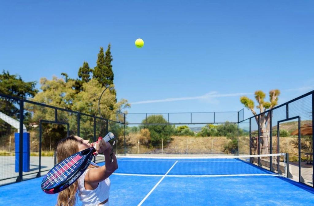 Jauna tenisiste gatavojas sitienam bumbu, kas lido gaisā, uz zila laukuma ar zaļo zāli fonā.