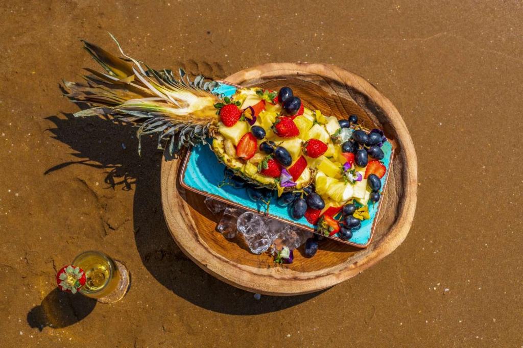 Ananāsa šķēle ar dažādiem augļiem, pasniegta uz zilas plāksnes, atrodas uz smiltīm pludmalē. Blakus novietota glāze ar dzērienu, rotāta ar zemeņu.