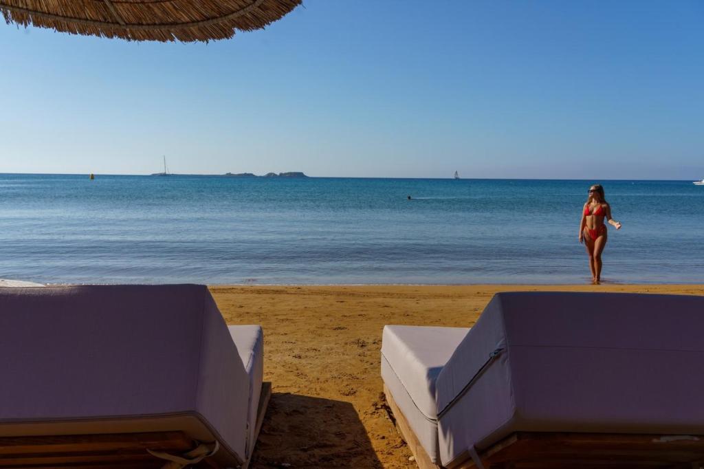 Pludmales skats ar smiltīm, sauli un mierīgu jūru, kur sieviete sarkanā peldkostīmā pastaigājas gar krastu, kamēr aiz viņas atrodas sauļošanās krēsli.