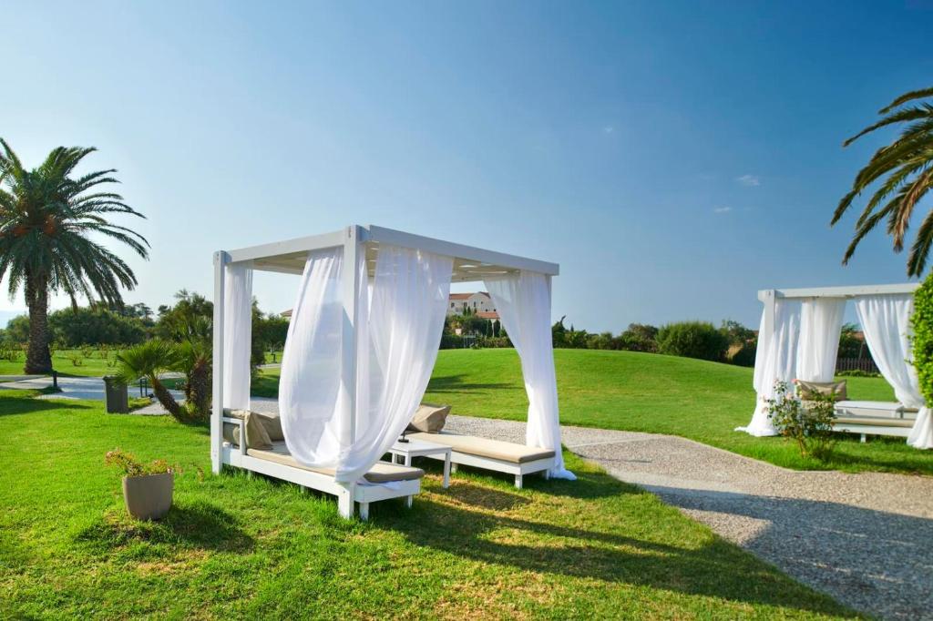 Balta atpūtas zona ar gultām un vieglām drānām, ierīkota zaļā parkā, apjozta ar palmu kokiem un skaidru debesu fonu.