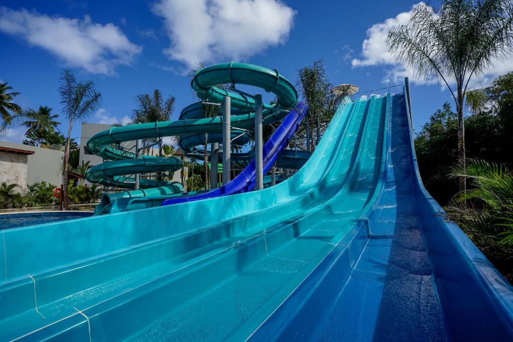 Ūdens slidkalniņi ar zilu virsmu un zaļiem elementiem, kas izvietoti baseina teritorijā ar palmām un skaidru debesu fonu. Ideāla vieta vasaras izklaidēm.