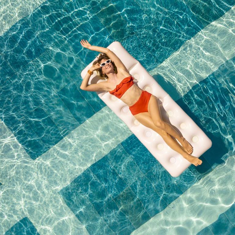 Sieviete oranžā peldkostīmā guļ uz peldošā matrača baseinā, baudot sauli un atpūtu. Ūdens ir dzidrs un mirdzošs, radot relaksējošu atmosfēru.
