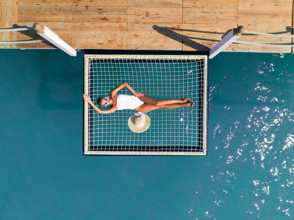 Sieviete baltā peldkostīmā atpūšas uz tīkla virs ūdens, valkājot sauļošanās brilles un cepuri. Fons ir dzidrs, zilas krāsas ūdens un koka laipa.