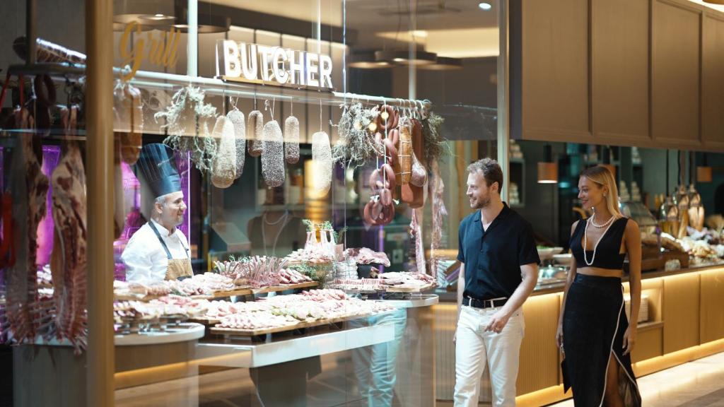 Svaigas gaļas veikals ar šefpavāru, kas pasniedz gaļas izstrādājumus, un divi apmeklētāji, kuri apskata piedāvājumu. Gaisīga un modernā vide, kas izraisa interesi par gaļas produktiem.