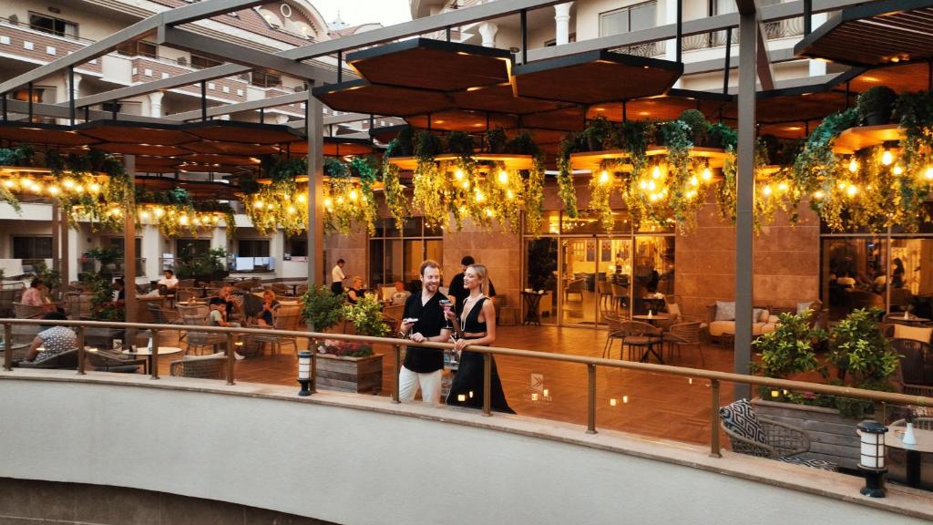 Eleganta terases aina ar zaļumiem un gaismiņām, kur pāris baudīja dzērienus. Fons ir rosīgs restorāns ar viesiem un mājīgu atmosfēru.