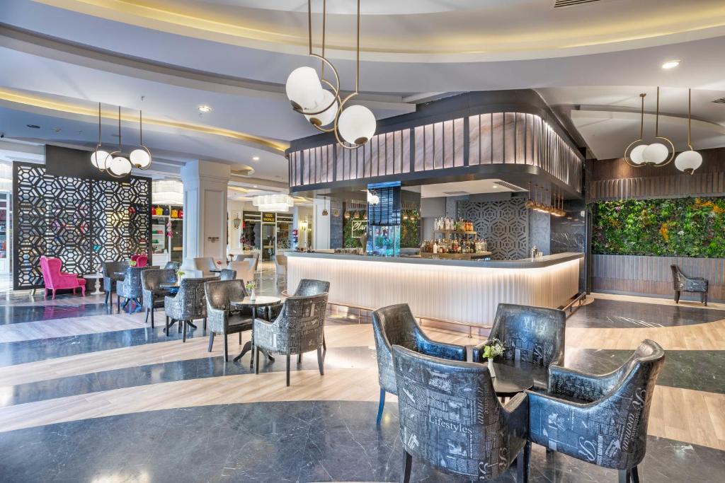 Mūsdienīgs viesnīcas bārs ar elegantu interjeru, kurā ir stilīgas krēslus un modernu apgaismojumu. Bāra zona izceļas ar dekoratīvām sienām un zaļumu, radot patīkamu atmosfēru.