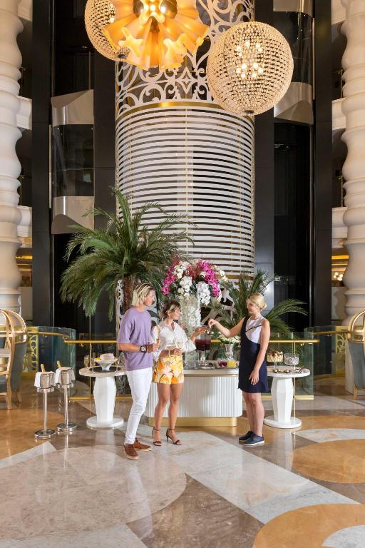 Elegants viesnīcas interjers ar viesmīlīgo apkalpošanu, kur divi apmeklētāji sarunājas pie greznā galda, kam blakus ir ziedu kompozīcija.