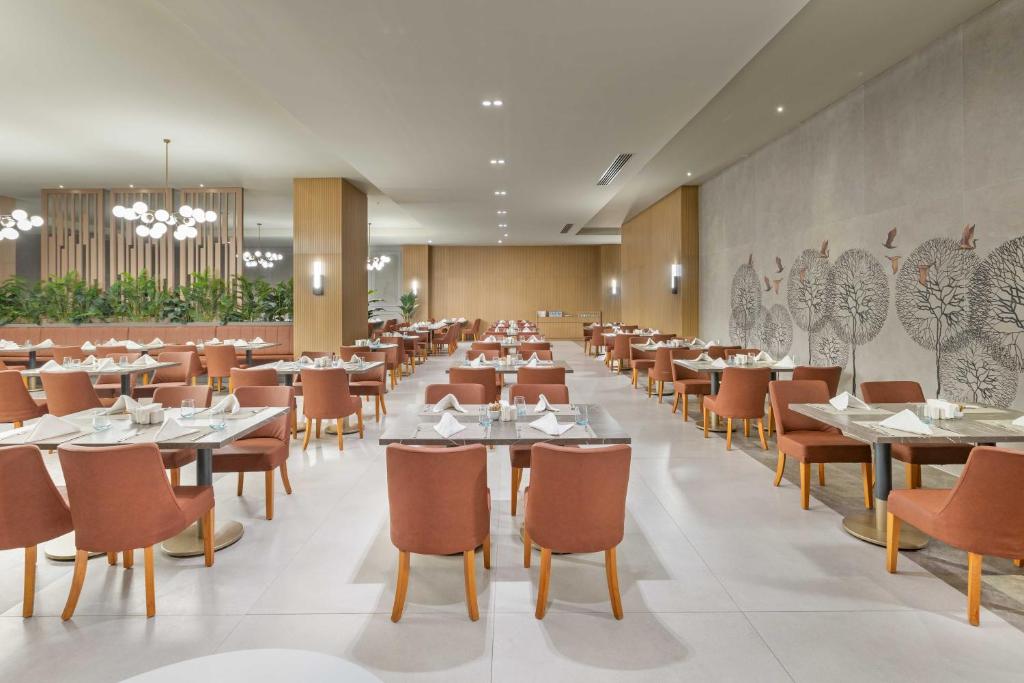 Mūsdienīgs restorāns ar elegantiem galdiem un krēsliem, kas izkārtoti plašā telpā, ar augstām griestiem un dekoratīviem elementiem. Svaigi zaļumi un stilizēti koki piešķir atmosfērai dzīvīgumu.