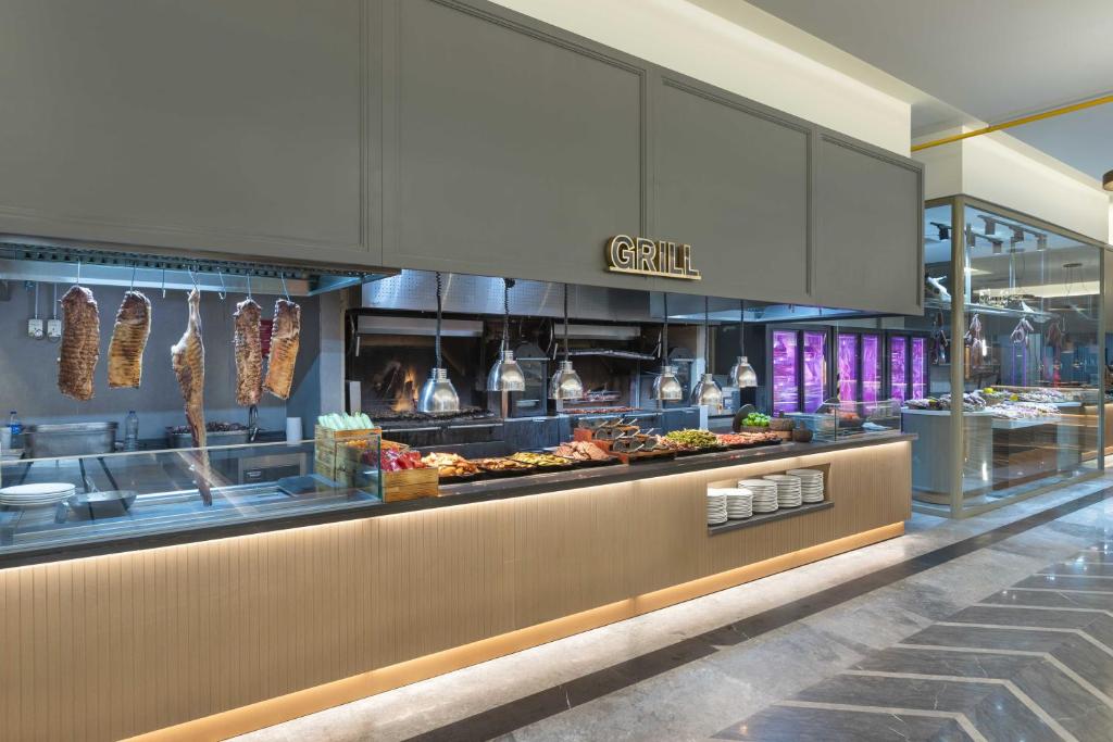 Grila restorāna skats ar gaļu, kas karājas uz āķiem, un dažādiem ēdieniem uz galda. Moderns interjers ar izgaismotām vitrīnām un grila zonu.