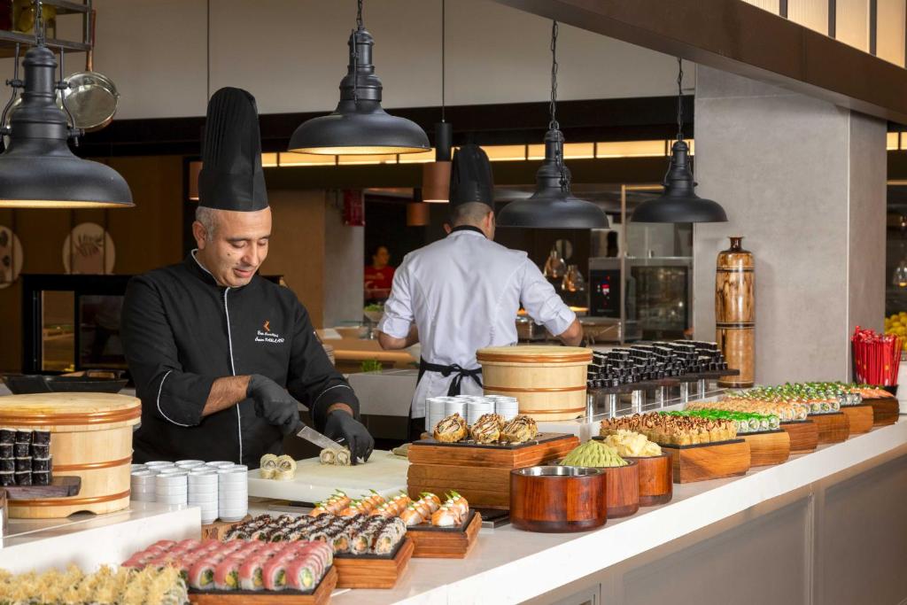 Šefpavārs gatavo suši dažādu veidu ēdienu bāzē, kurā redzami krāsaini suši un citi japāņu virtuves ēdieni. Svaigi pagatavotas delikateses ir sakārtotas uz koka traukiem, radot pievilcīgu skatu.