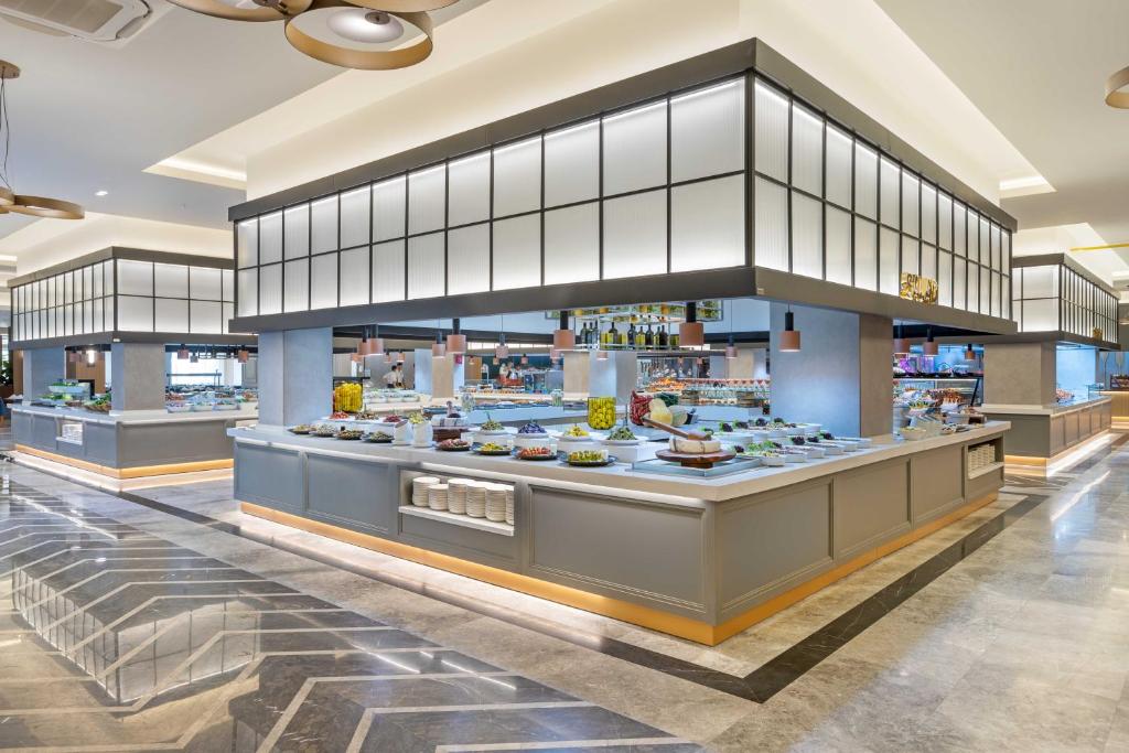 Mūsdienīga bufete ar elegantiem galdiem, kas pilni ar dažādiem ēdieniem un dzērieniem, izgaismota ar siltu apgaismojumu. Stilīgs interjers ar marmora grīdu un modernām dizaina detaļām.