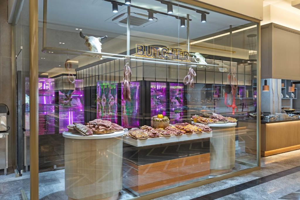 Mūsdienīgs gaļas veikals ar plašu gaļas produktu klāstu, izstādīts uz baltiem galdiem un iepriekš atdzisēts vitrīnās. Aiz stikla sienas redzams spilgti apgaismots veikals ar stilīgu interjeru.