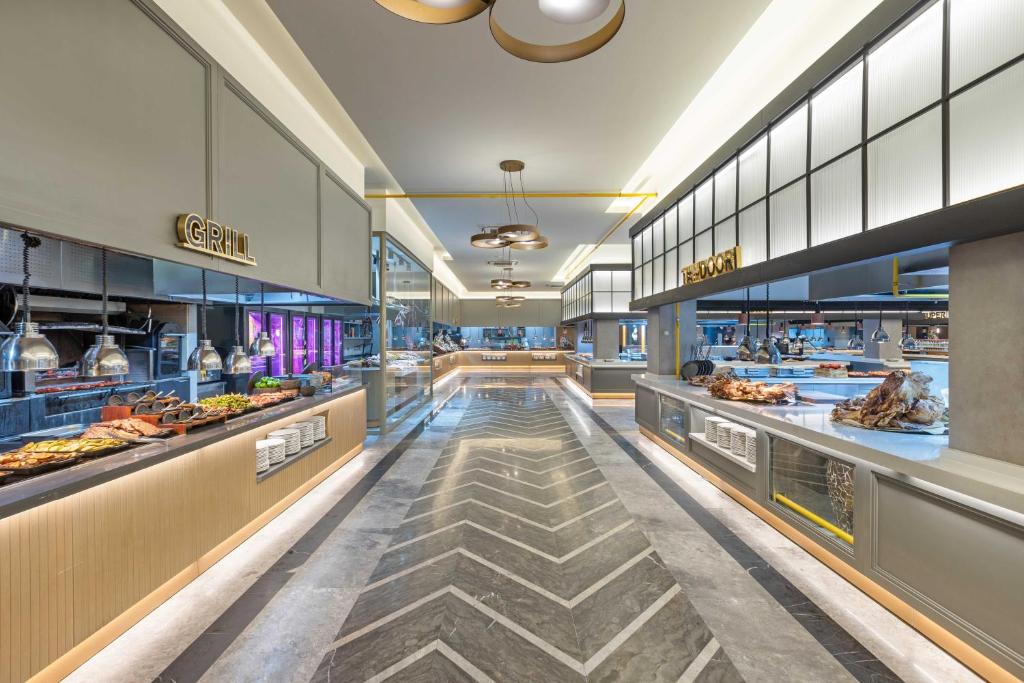 Mūsdienīgs restorāns ar elegantu interjeru, kurā redzamas grila un citu ēdienu gatavošanas zonas. Apgaismojums un marķējums piešķir telpai stilīgu atmosfēru.