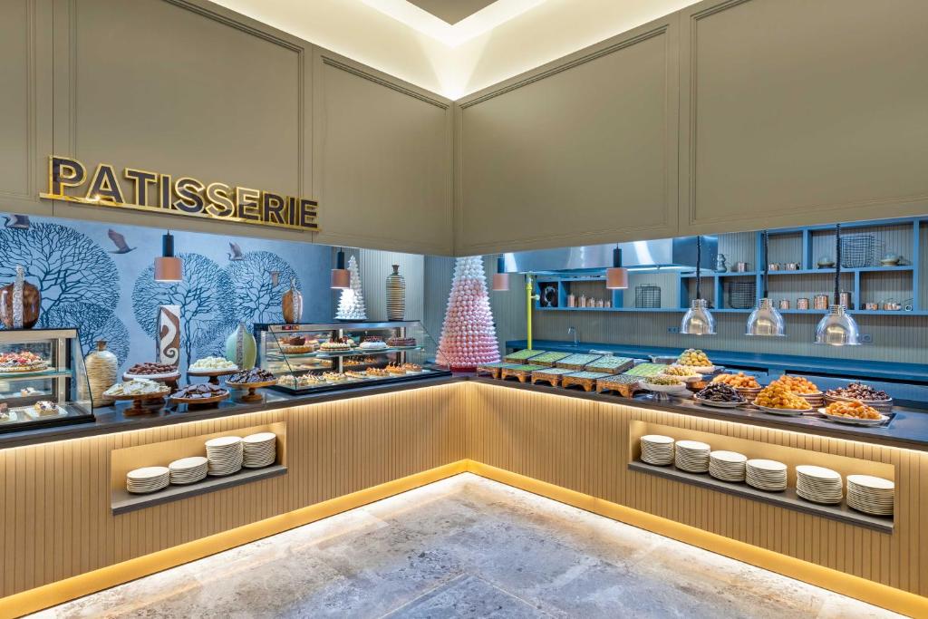 Saldumu veikals ar elegantiem desertiem un modernu interjeru, kurā redzamas dažādas kūkas un uzkodas. Gaisma un dizains rada siltu un pievilcīgu atmosfēru.