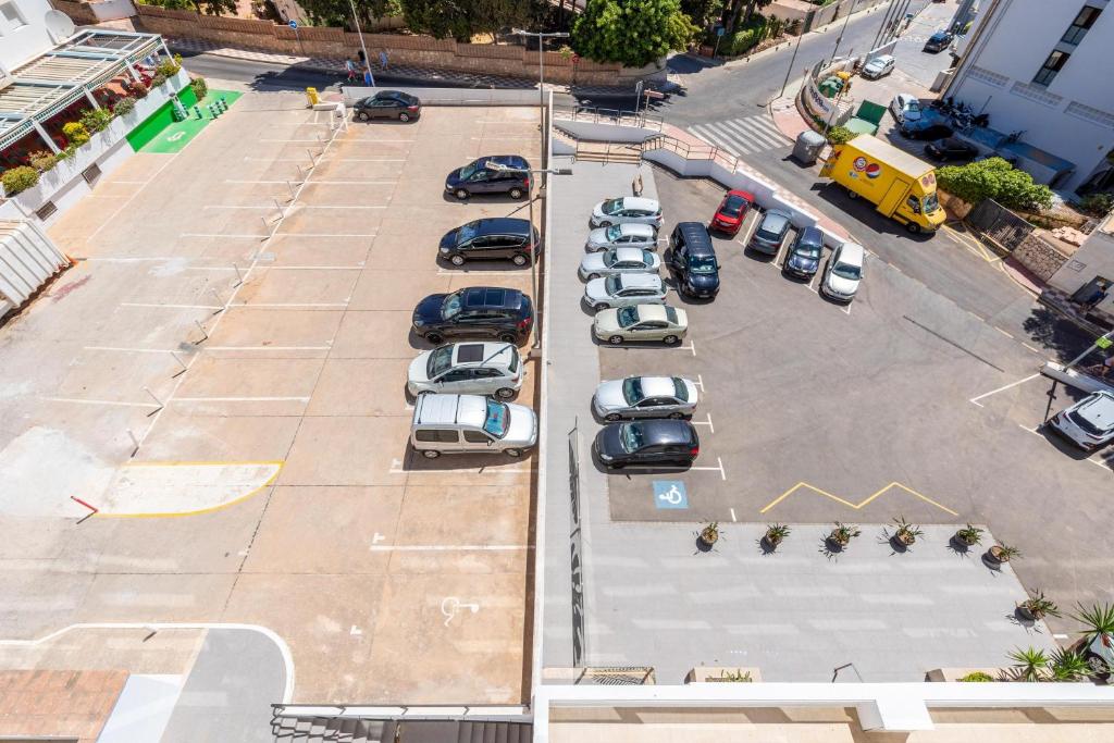 Autostāvvieta ar dažādiem automobiļiem un brīvām vietām, redzama no augšas. Stāvvieta ir sadalīta divās daļās, un blakus ir ēka un ceļš.