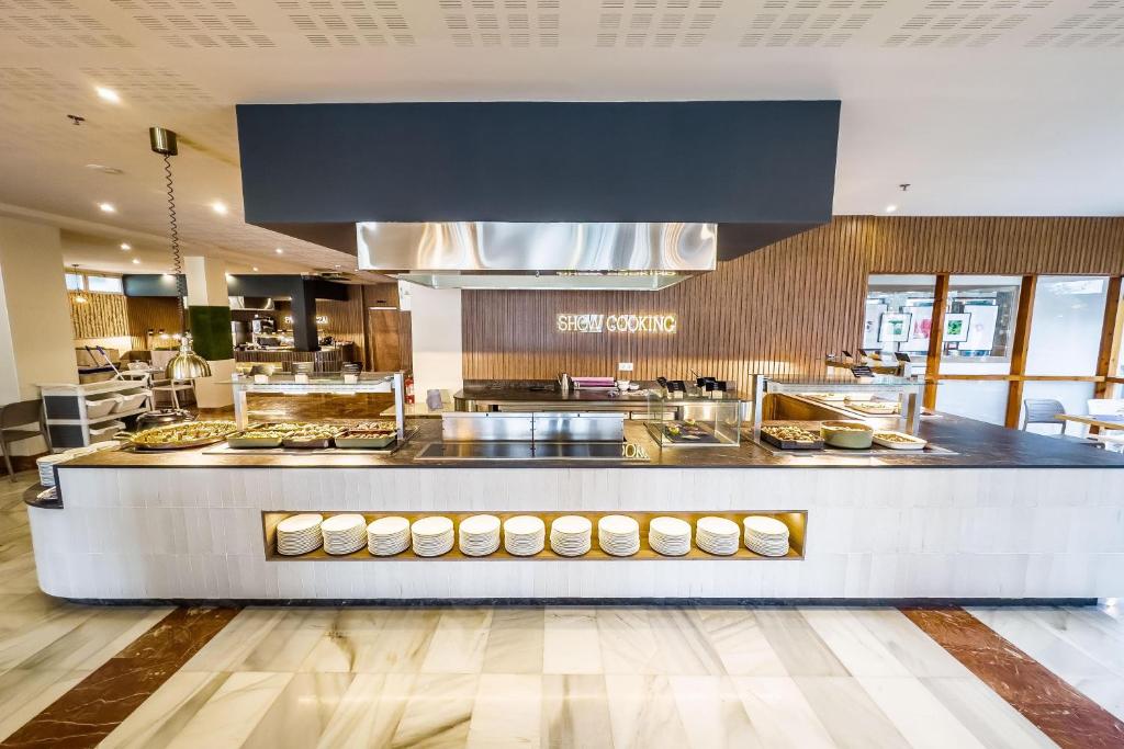 Mūsdienīga restorāna zona ar atvērtu virtuvi un pašapkalpošanās bāru, kurā redzama ēdienu gatavošana un plašs galdu klāsts. Interjers ar koka akcentiem un elegantu apgaismojumu.