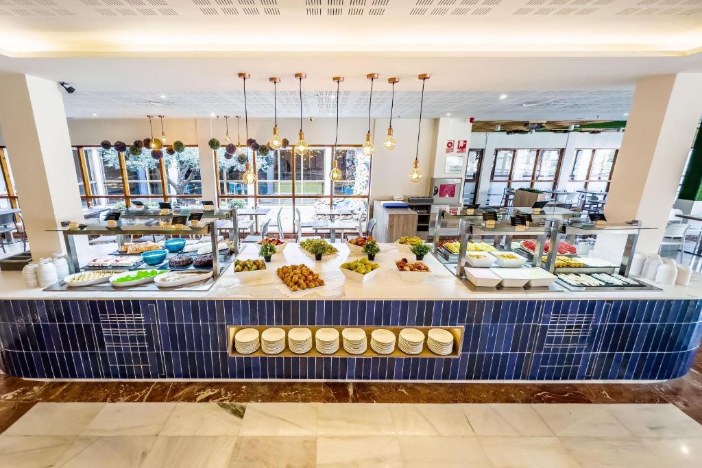 Labi iekārtota bufete ar dažādiem ēdieniem un svaigiem augļiem, apgaismota ar stilīgām gaismas ķermeņiem. Interjers ir gaišs un moderns, radot patīkamu atmosfēru.