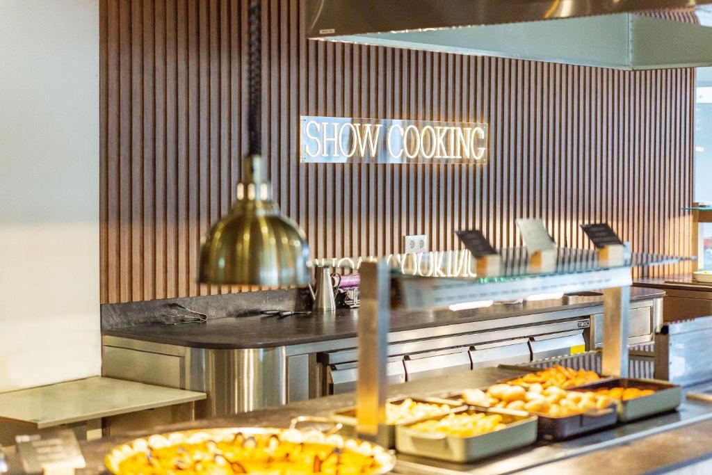 Mūsdienīga restorāna virtuve ar "Show Cooking" zīmi, kur redzams dažādu ēdienu izvēle uz galda. Elegantais interjers un apgaismojums rada patīkamu atmosfēru.